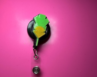 Puede incluir: Un carrete de insignia retráctil negro con un adorno en forma de hoja de color verde brillante y amarillo. El carrete está unido a una correa de plástico transparente con un botón de metal, conectado a un porta credenciales blanco.
