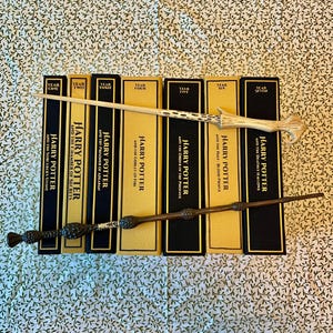 May include: A collection of seven Harry Potter books with matching wands. The books are in a row, with gold spines and black covers. The wands are light brown and dark brown, with intricate designs.