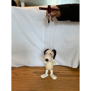 May include: A Snoopy puppet with a wooden body, black ears, and a black spot on top of his head. The puppet is suspended by red strings attached to a wooden control bar. The puppet's arms are outstretched.