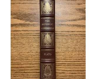 Puede incluir: Un lomo de libro de color marrón oscuro con letras en pan de oro y detalles decorativos. El texto dice THE REPUBLIC y PLATO. Tres paneles rectangulares presentan diseños de palmetas doradas. El lomo tiene bandas horizontales en relieve.