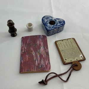 Könnte beinhalten: Eine Sammlung von Gegenständen, darunter ein dunkler Metallstreuer, ein kleiner weißer Keramikbecher, eine herzförmige blau-weiße Keramikdose, ein Notizbuch mit marmoriertem kastanienbraunem Einband und eine hölzerne Fibel mit einer Messingplatte.