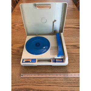 May include: A beige and blue Fisher-Price toy record player with a hinged lid, a blue circular turntable, and a blue tone arm. A metal ruler placed in front shows the device is approximately 16 inches wide. The unit features volume controls.