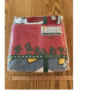 May include: A Disney Store Santa's Workshop handmade quilted throw in plastic packaging. The label features red, white, and green colors with silhouettes of cartoon characters. A wooden ruler shows the item is approximately 15 inches wide.