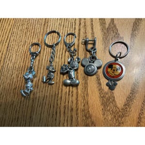 Op de afbeelding: Vijf metalen sleutelhangers met Mickey Mouse-ontwerpen. Vier zijn zilverkleurige figuren of vormen, waaronder een staand personage, een personage met een ster en een kompas. De vijfde is een ronde medaillon met een rood midden en de tekst Mickey Mouse Club.