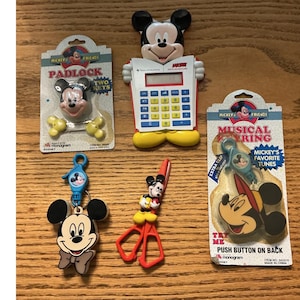 May include: A collection of five Disney Mickey Mouse themed items including a padlock with two keys, a Texas Instruments calculator, a keychain, a pair of red scissors, and a musical keyring, all featuring Mickey Mouse character designs and branding.