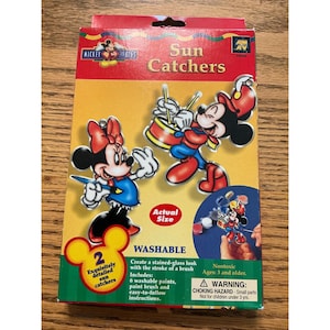 Op de afbeelding: Een doosje met zonnevangers met Mickey en Minnie Mouse. De doos is rood, geel en groen, met de woorden "Sun Catchers" en "Washable" erop gedrukt. De set bevat 6 wasbare verven, een penseel en instructies.
