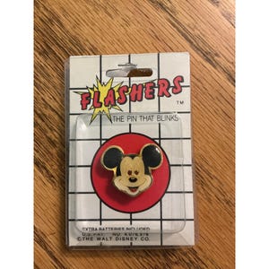 May include: A vintage Flashers brand pin featuring a gold-toned Mickey Mouse head with black ears, set against a red circular background on a white grid-patterned backing card. The packaging includes the text Flashers, The Pin That Blinks.