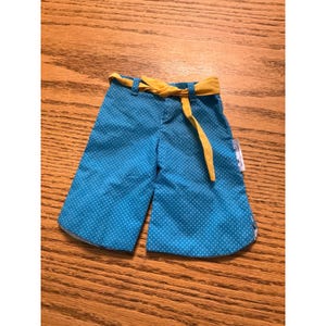 May include: A pair of bright blue wide-leg pants featuring a small white polka dot pattern throughout. The pants include belt loops and a mustard yellow fabric tie belt threaded through the loops at the waist. A small white tag is visible on the side.