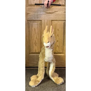 Può includere: Un pupazzo di coniglio marrone chiaro con orecchie e arti lunghi. Il pupazzo è controllato da una barra nera. Il coniglio ha un petto bianco e un'espressione sorridente.