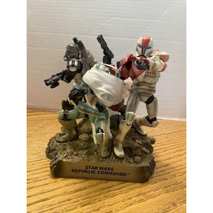 Puede incluir: Figura detallada con tres Star Wars Republic Commandos en poses dinámicas. Las figuras visten armaduras blancas, rojas, verdes y negras. La base de la figura tiene el texto "STAR WARS REPUBLIC COMMANDO™".
