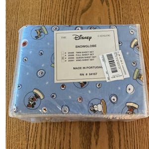 Puede incluir: Un juego de sábanas azul claro en embalaje de plástico con un patrón repetitivo de bolas de nieve de Disney con personajes y muñecos de nieve. Una etiqueta blanca dice THE DISNEY CATALOG SNOWGLOBE, Made in Portugal, RN # 84167.