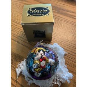 Puede incluir: Un adorno navideño de cristal redondo con un personaje de Mickey Mouse pintado con sombrero amarillo y pajarita roja, sobre un fondo azul oscuro y morado con estrellas brillantes. Se acompaña de una caja dorada de la Polonaise Collection.