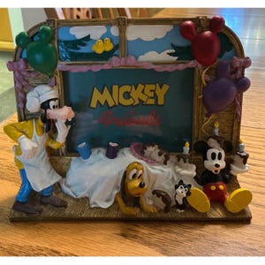 Puede incluir: Un marco de fotos decorativo con Mickey Mouse y sus amigos. El marco tiene un fondo verde azulado con las palabras "Mickey Friends". El marco incluye imágenes de Goofy, Pluto, Mickey Mouse y otros personajes. El marco está decorado con globos y un pastel.
