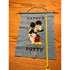 May include: A light blue felt wall hanging featuring a graphic of a cartoon mouse sitting on a toilet with the text PATSY'S POTTY. A yellow measuring tape shows the vertical length is approximately 22 inches from the top rod to the bottom edge.