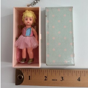 May include: A small doll with blonde hair, wearing a pink and blue bodice with a pink tulle skirt, inside a light pink box next to a light blue box with pink polka dots. A metal ruler shows the doll and boxes are approximately 3.5 inches tall.