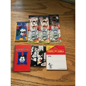 May include: A collection of Mickey Mouse stationery items including three packages of jumbo paper clips, a spiral-bound notebook with a red cover, a small black and white character notebook, and a pad of Post-it Notes featuring a small Mickey Mouse graphic.