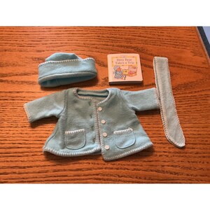 Peut inclure: Une veste en polaire bleu clair avec des coutures blanches, deux poches avant et quatre boutons blancs. L'ensemble comprend un bonnet bleu clair assorti, une longue écharpe bleu clair avec des coutures blanches et un petit livre Bitty Bear Takes a Trip.