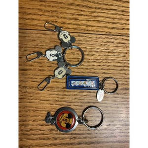 Könnte beinhalten: Drei Metall-Schlüsselanhänger mit Disney-Figuren. Ein Anhänger hat zwei Mickey-Mouse-Kopf-Charms, ein weiterer ist ein rechteckiger blauer Rahmen mit einer grafischen Szene und der dritte ist ein rundes, rot-schwarzes Mickey-Mouse-Club-Logo.