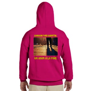 May include: A magenta hoodie with a hood, featuring a yellow-bordered image on the back. The image shows a person walking on a sunlit street. Above the image, the text reads "CHAQUE PAS COMPTE." Below the image, the text reads "UN JOUR À LA FOIS."