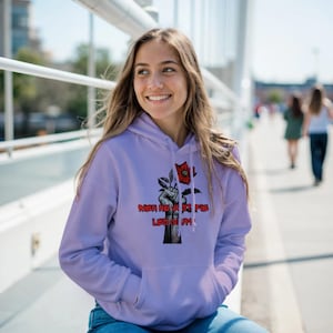 May include: A lilac hoodie featuring a graphic of a clenched fist holding a flower, with the text "MEN NE SONT FIE LEGION!". The hoodie has a kangaroo pocket and a drawstring hood.