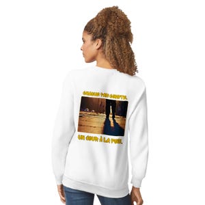 May include: White sweatshirt with a graphic on the back featuring a person walking on a city street at night. The graphic includes the text "CHAQUE PAS COMPTE" and "UN JOUR À LA FOIS" in yellow.