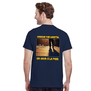 May include: Navy blue t-shirt with a graphic on the back featuring a person walking on a city street. The text "CHAQUE PAS COMPTE. UN JOUR À LA FOIS." is printed above and below the image in yellow.