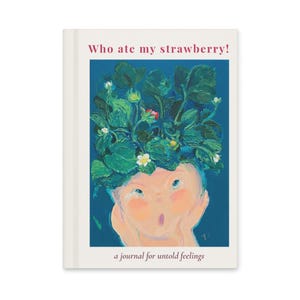 May include: A journal with a cream-colored cover featuring an illustration. The illustration depicts a face with a strawberry plant for hair, the text "Who ate my strawberry!" and the text "a journal for untold feelings".