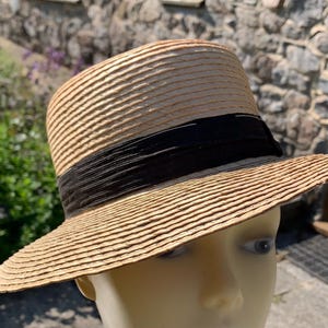 May include: A straw boater hat with a black grosgrain ribbon band. The hat has a flat crown and a wide, slightly curved brim. The hat is made of woven straw and is a light tan color.
