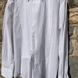 May include: A white dress shirt with a pleated bib front and a classic collar. The long-sleeved shirt has buttoned cuffs and a button-down front. The shirt is hanging against a stone wall.