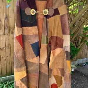 May include: A patchwork suede cape with a brown, red, and green color scheme. The cape has a collar and is fastened with a gold chain and two circular clasps.