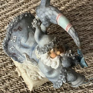 Puede incluir: Una figura caprichosa de una figura sentada con un atuendo azul con barba y sombrero blancos, sosteniendo un pequeño pájaro azul. La figura está sobre una base con notas musicales y un pájaro posado encima. La figura tiene una combinación de colores azul claro, blanco y melocotón.