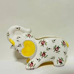 Puede incluir: Jardinera de cerámica blanca con forma de elefante, con detalles amarillos y un estampado floral de rosas rojas y hojas verdes. El elefante tiene la trompa levantada y la parte superior abierta.