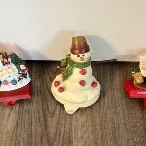 Puede incluir: Tres sujetadores de calcetines navideños. Uno presenta a Papá Noel, otro un muñeco de nieve con una bufanda verde y manzanas rojas, y el tercero representa niños y regalos. Todos están sobre bases rojas.