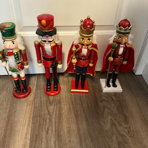 May include: Four decorative wooden nutcrackers in various designs. The nutcrackers are dressed in red, gold, and green outfits, with hats and crowns. One nutcracker holds a staff. The nutcrackers stand on a wooden floor.