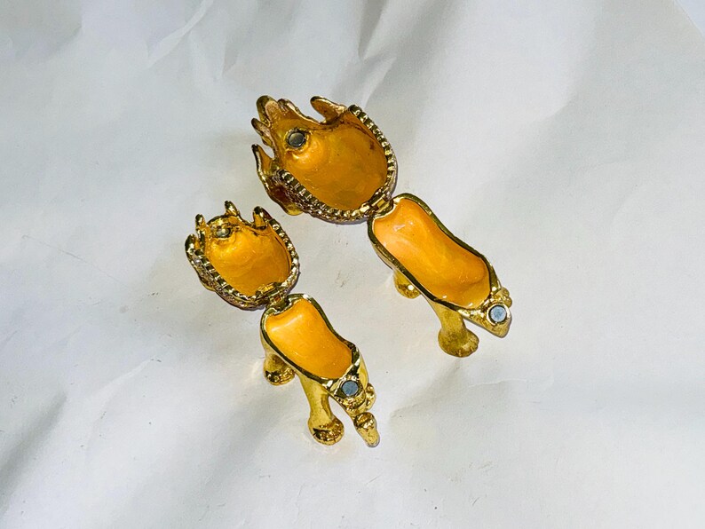 Puede incluir: Dos joyeros dorados con forma de zapatos de tac&oacute;n alto, con un interior amarillo brillante y peque&ntilde;as joyas. Las cajas con bisagras est&aacute;n abiertas, revelando el interior. Los zapatos est&aacute;n sobre una superficie blanca.