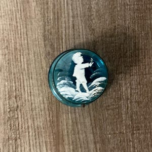 Puede incluir: Un objeto redondo de vidrio de color turquesa con una silueta blanca de una persona en un campo. La persona está extendiendo la mano hacia el cielo. El objeto está sobre un fondo de madera.