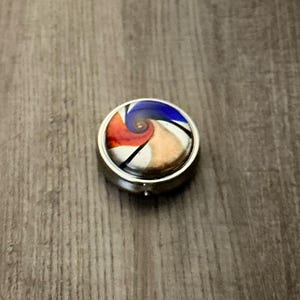 Può includere: Un piccolo portapillole rotondo argentato con un design astratto colorato sul coperchio. Il design presenta un motivo a spirale con tonalità rosse, blu e beige. Il portapillole è posto su una superficie di legno.