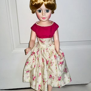 May include: A vintage doll with blonde hair styled in an updo. She wears a pink top with a scalloped neckline and a cream-colored dress adorned with pink roses. The doll is wearing clear heels.
