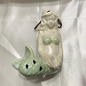 Pode incluir: Estatueta de sereia de cerâmica com acabamento perolado. A sereia tem um sutiã em forma de concha verde e uma cauda verde clara com recortes em forma de diamante. Uma corda marrom é presa ao topo da cabeça.