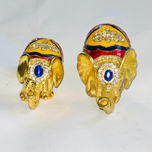 Puede incluir: Dos figuras de elefantes dorados y ornamentados. Cada elefante tiene un tocado enjoyado amarillo y rojo, una joya azul en el centro y est&aacute; incrustado con pedrer&iacute;a transparente. Probablemente son art&iacute;culos decorativos o joyeros.
