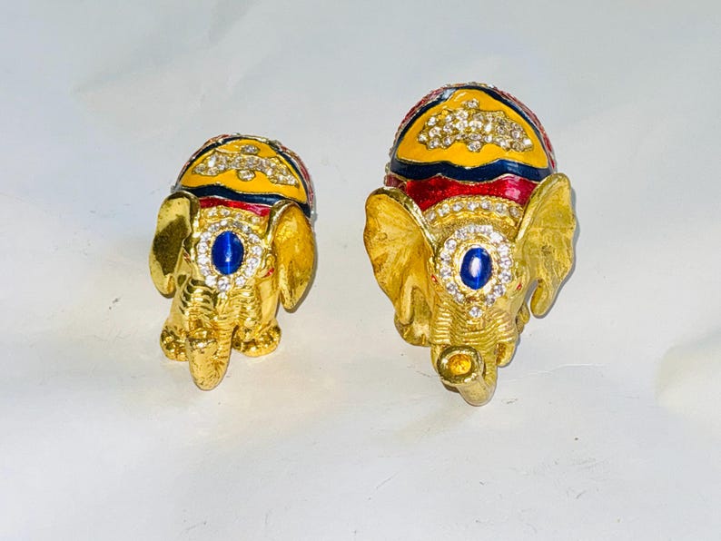 Puede incluir: Dos figuras de elefantes dorados y ornamentados. Cada elefante tiene un tocado enjoyado amarillo y rojo, una joya azul en el centro y est&aacute; incrustado con pedrer&iacute;a transparente. Probablemente son art&iacute;culos decorativos o joyeros.