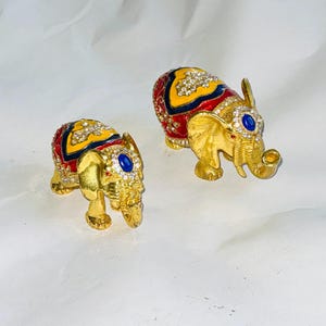 Puede incluir: Dos figuras de elefantes dorados. Cada elefante tiene un dise&ntilde;o estampado en rojo, amarillo y azul en la espalda, adornado con pedrer&iacute;a transparente. Una gema azul est&aacute; engastada en la frente de cada elefante.