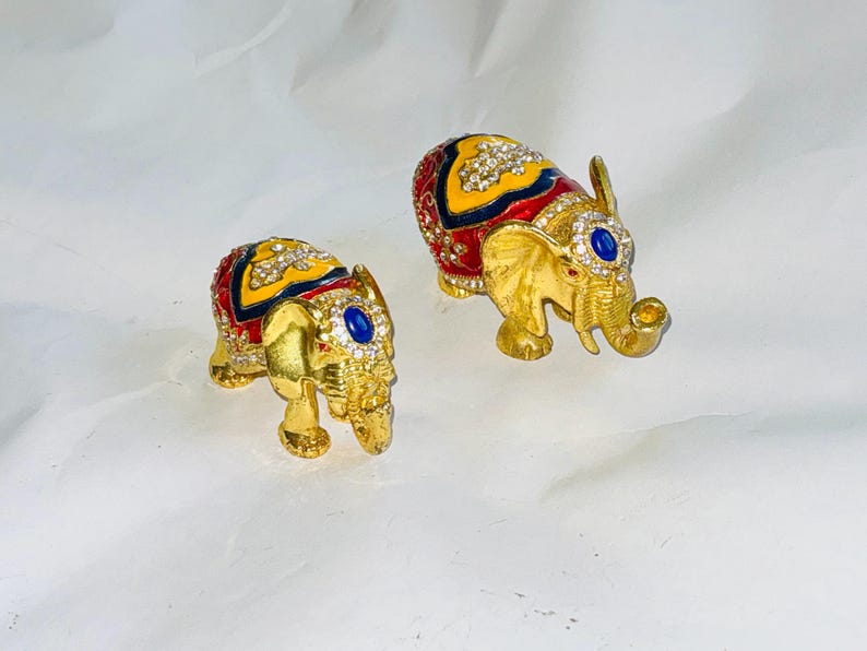 Puede incluir: Dos figuras de elefantes dorados. Cada elefante tiene un dise&ntilde;o estampado en rojo, amarillo y azul en la espalda, adornado con pedrer&iacute;a transparente. Una gema azul est&aacute; engastada en la frente de cada elefante.