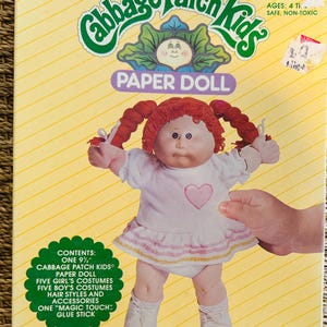 May include: A vintage Cabbage Patch Kids Paper Doll box. The box features a doll with red pigtails, wearing a white dress with a pink heart. The box is yellow with the words "Cabbage Patch Kids" and "Paper Doll". The box includes a 9.5-inch doll, costumes, and accessories.