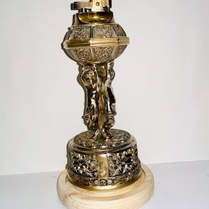 May include: A vintage brass and silver-toned lighter with intricate detailing. The lighter features a globe-shaped body supported by sculpted figures, mounted on a wooden base. The top portion is gold-toned, and the base is silver-toned.