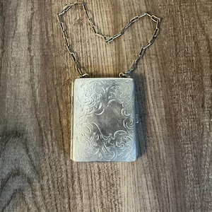 May include: Silver rectangular case with ornate floral engraving and a chain handle. The case is closed and appears to be made of metal. The background is a wooden surface.