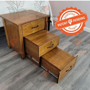 May include: A wooden pet step with three drawers, stained in a warm brown hue. Each drawer has a black handle. The steps are designed to help pets reach higher places. The image includes a "Patent Pending" seal.