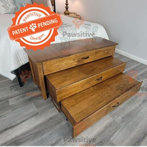 May include: A three-tiered wooden pet step with pull-out drawers. The steps are a warm brown color with black handles. The top step has a "Patent Pending" sticker. The steps are placed next to a bed.