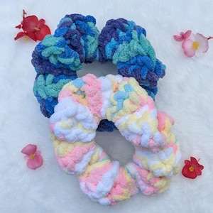 May include: Two handmade scrunchies in the image. One is a mix of blue and teal, the other is pastel with pink, yellow, and white. The scrunchies are made of a soft, textured yarn. Decorative flowers are scattered around the scrunchies.