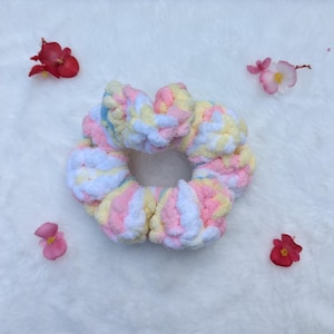 May include: A handmade, pastel-colored scrunchie with a soft, textured appearance. The scrunchie features a mix of pink, yellow, blue, and white yarn. The background is a white, fluffy surface, with scattered red and pink flower petals.